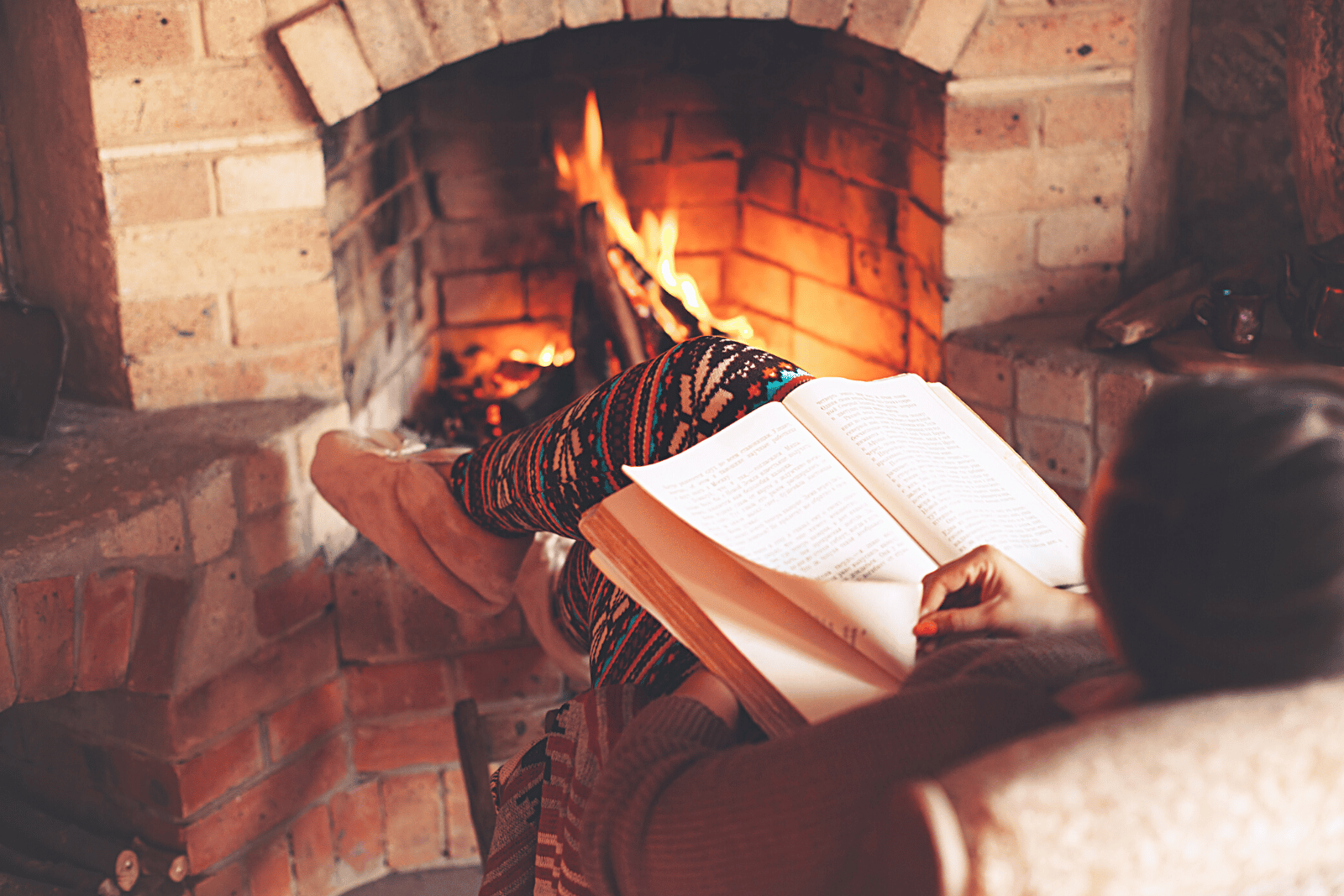 Image featuring a woman with Christmas pajamas on her, reading in a book, sitting in front of a cozy fireplace with a warm glow illuminating the room