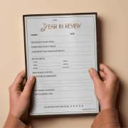 Hands hold a "Year In Review" worksheet on beige background. The form includes sections for highlights, favorites, memories, and a year rating.